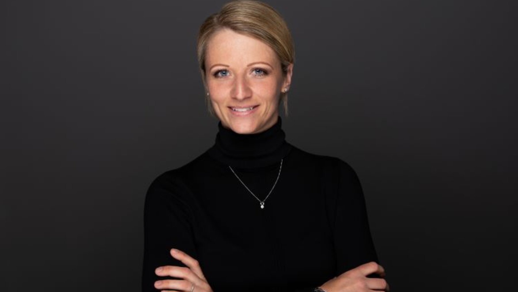 A woman with short blonde hair is standing with her arms crossed. She is wearing a black turtleneck top and a necklace. The background is plain and dark, putting the focus on her. She has a slight smile on her face.