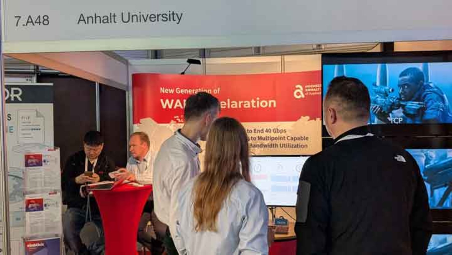 Messebesucher stehen vor einem Stand der Anhalt Universität mit dem Thema „New Generation of WLAN Acceleration“. Ein großer Bildschirm zeigt zwei Personen bei der Arbeit mit Technik. Drei Personen unterhalten sich, eine weitere Person sitzt am Stand und notiert etwas.
