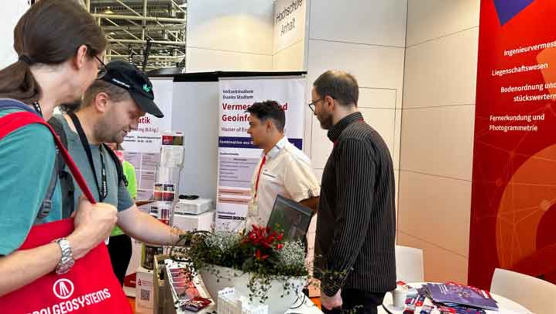 Drei Personen stehen an einem Messestand mit dem Thema Vermessung und Geoinformatik. Ein Mann in rot-weißer Jacke und ein anderer in schwarzem Hemd unterhalten sich mit einer Frau in grüner Jacke, die sich über eine Tischauslage mit Prospekten und einer kleinen Pflanze mit roten Blüten beugt.