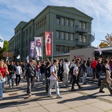 Große Gruppe von Studierenden marschiert vor einem grünen Hochschulgebäude mit dem Schriftzug Hochschule Anhalt. Sie tragen Plakate, Fahnen und Transparente mit der Zahl 6 und dem Logo der Hochschule. Es ist eine Führung für die neuen Studierenden zur Orientierung.