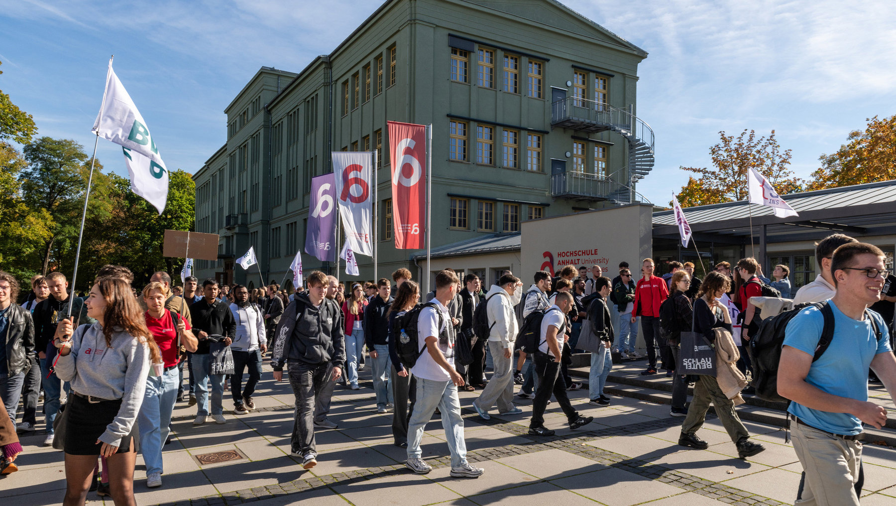 Große Gruppe von Studierenden marschiert vor einem grünen Hochschulgebäude mit dem Schriftzug Hochschule Anhalt. Sie tragen Plakate, Fahnen und Transparente mit der Zahl 6 und dem Logo der Hochschule. Es ist eine Führung für die neuen Studierenden zur Orientierung.