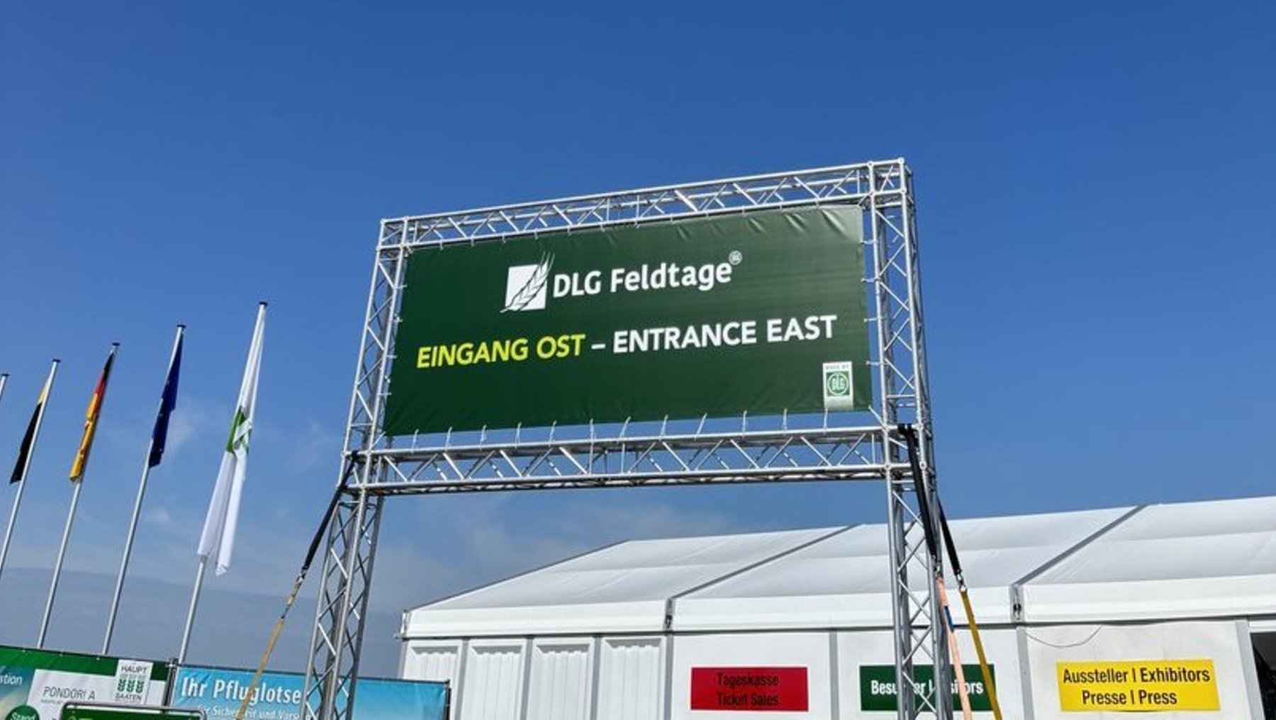 Grüner Schilderaufbau mit weißer Schrift: „DLG Feldtage“ und „Eingang Ost – Entrance East“ vor blauem Himmel, markiert den Ost-Eingang einer Messe oder Veranstaltung.