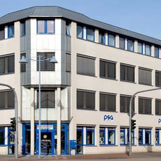 A three-story building with a flat roof. The ground floor houses a bank (Deutsche Bank) and a PSG office. The building has a modern design with large windows and a light-colored facade. The street in front has traffic lights and a no-parking sign.