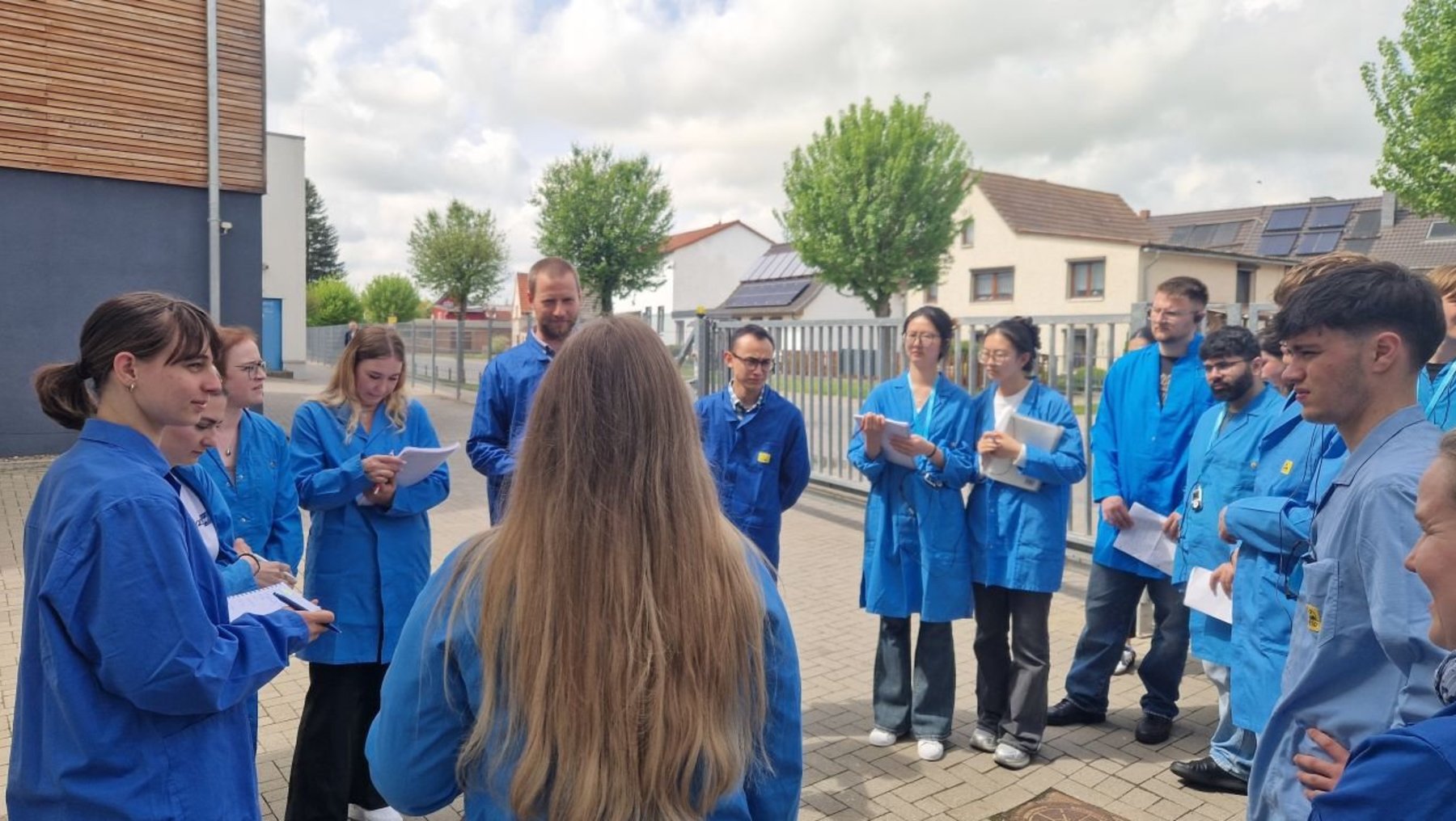 Gruppe von Menschen in blauen Arbeitskleidern steht im Freien und hört einer Person mit langem Haar zu, die ihnen etwas erklärt. Alle halten Notizblätter oder -blöcke und scheinen aufmerksam zuzuhören.