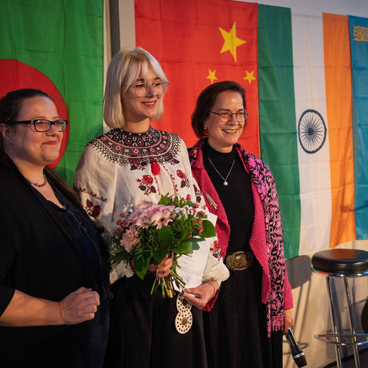 Drei lächelnde Frauen stehen vor einer Wand mit verschiedenen Flaggen, darunter die von Irland, China, Indien und Kasachstan. Die Frau in der Mitte hält einen Blumenstrauß und ein Dokument, während links und rechts Gitarren zu sehen sind.