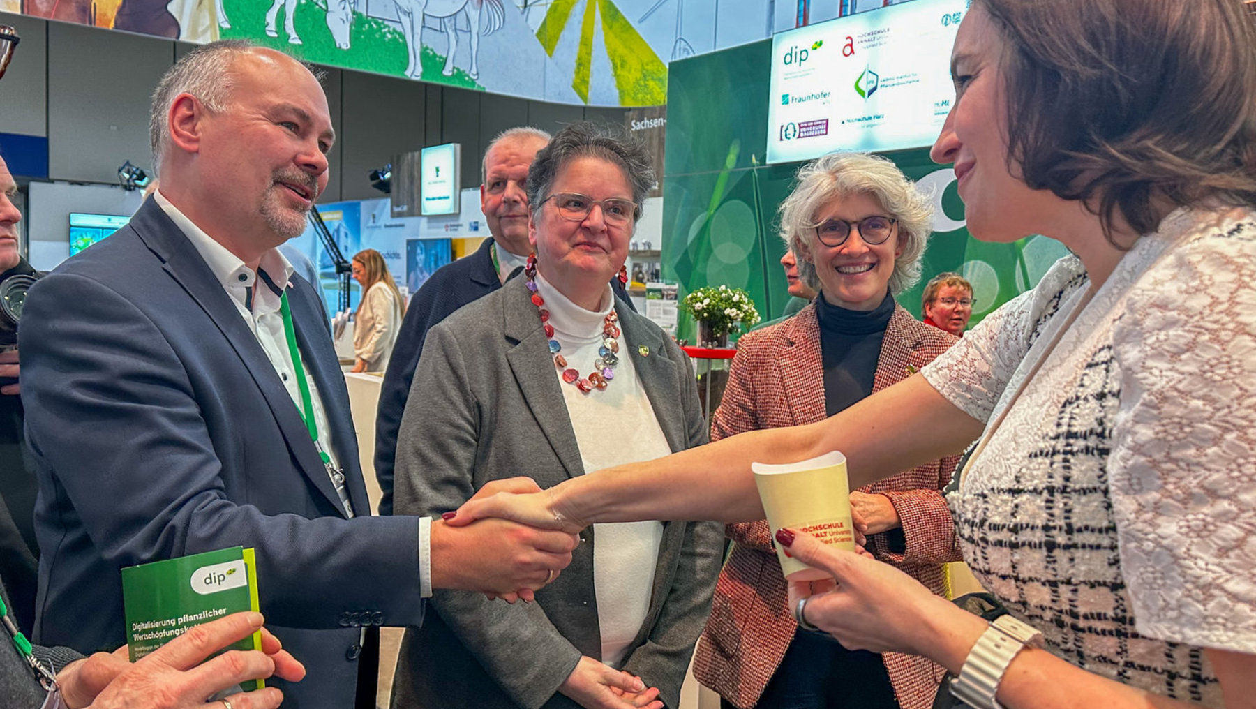 Ein Mann und eine Frau auf einer Messe reichen sich freundlich einen Pappbecher mit der Aufschrift „HOCHSCHULE“ überreichend. Im Hintergrund sind weitere Personen und ein Stand mit grünem Design und Logos wie „dip“ und „Fraunhofer“ zu sehen.