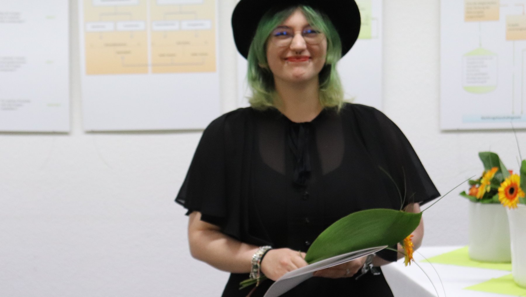 Eine Person mit grünen Haaren und schwarzem Kleid, darunter einer schwarzen Kapuze, hält ein Diplom oder eine Urkunde in den Händen und lächelt zufrieden. Im Hintergrund sind Informationsplakate an einer Wand befestigt.