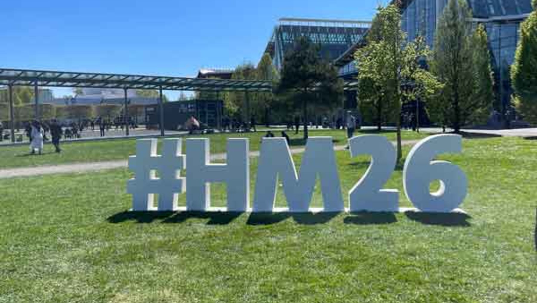 Großer Schriftzug auf Rasenfläche mit dem Hashtag und der Nummer **#Hm26**, umgeben von einem modernen Gebäudekomplex mit Glasdach und Menschen im Hintergrund.