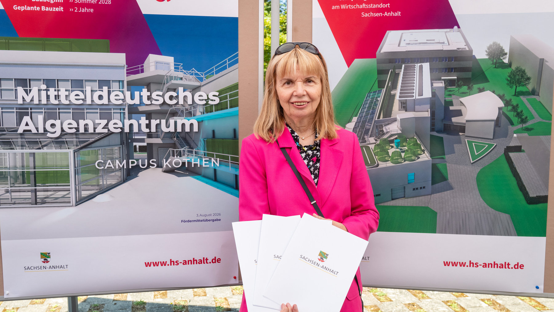 Eine lächelnde Person hält vor einem Plakat mit dem Titel „Mitteldeutsches Algenzentrum, Campus Köthen“ zwei Dokumente. Das Plakat zeigt Baupläne und Informationen zur Förderung des Projekts durch Sachsen-Anhalt.