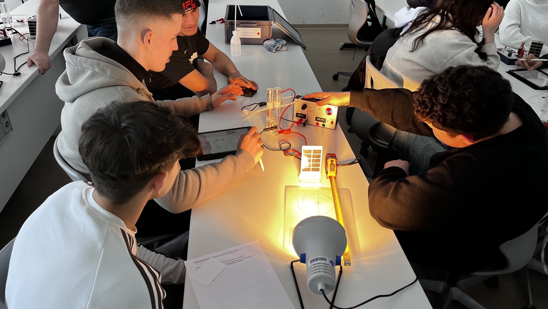 Eine Gruppe von Personen arbeitet gemeinsam an einem Tisch mit einer einfachen elektrischen Schaltung, die eine Glühbirne zum Leuchten bringt. Drähte sind mit einer Steckplatine verbunden, während alle konzentriert an der Funktionsweise experimentieren.