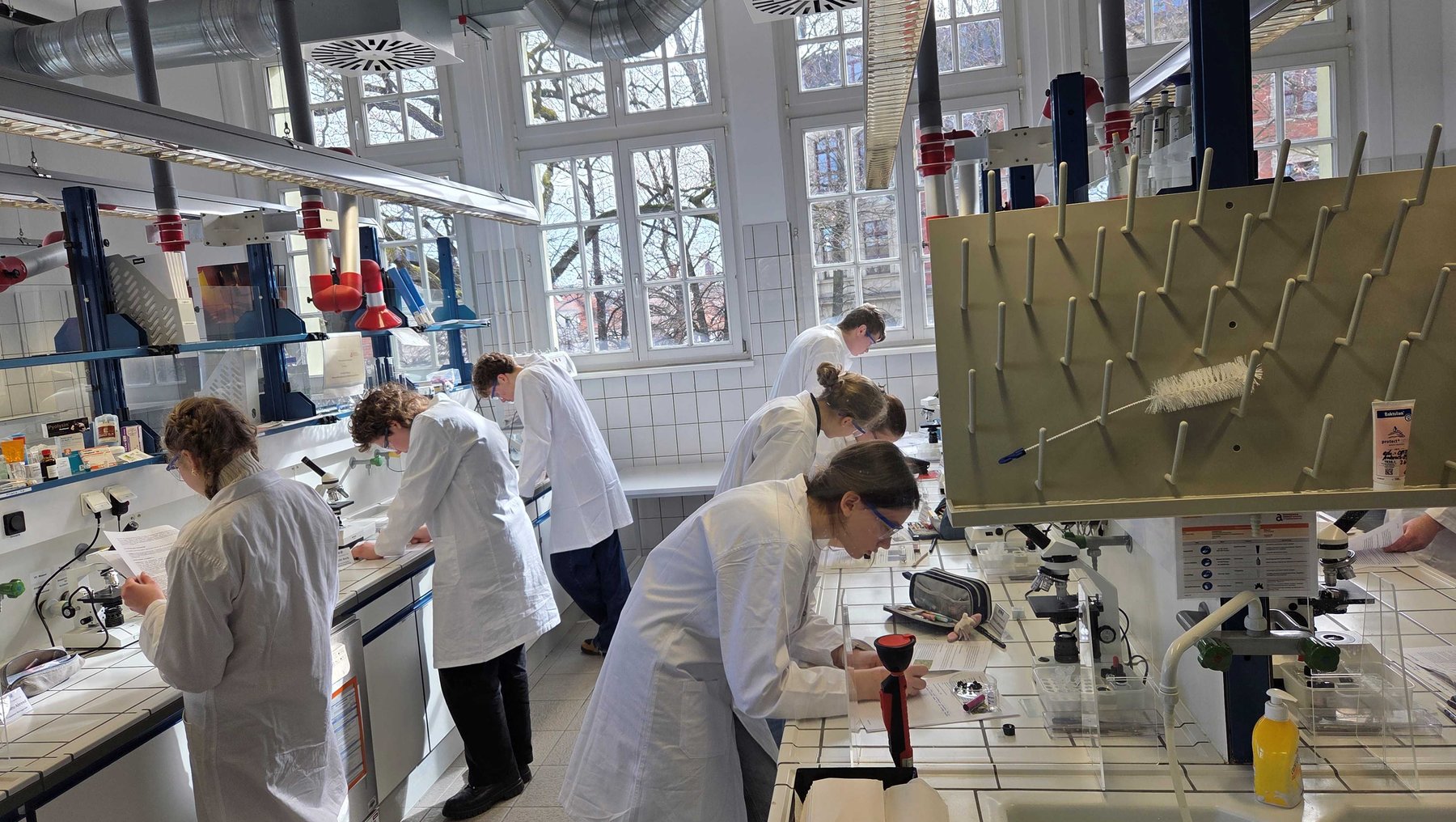 Drei Personen in weißer Laborkleidung arbeiten in einem hellen Labor: Zwei stehen an Arbeitsplätzen mit Chemikalien und Geräten, eine dritte bearbeitet vorsichtig eine Probe an einem Tisch mit Abzugshaube. Im Hintergrund sind weitere Laborutensilien und ein großes metallisches Gerüst mit Rillen zu sehen.