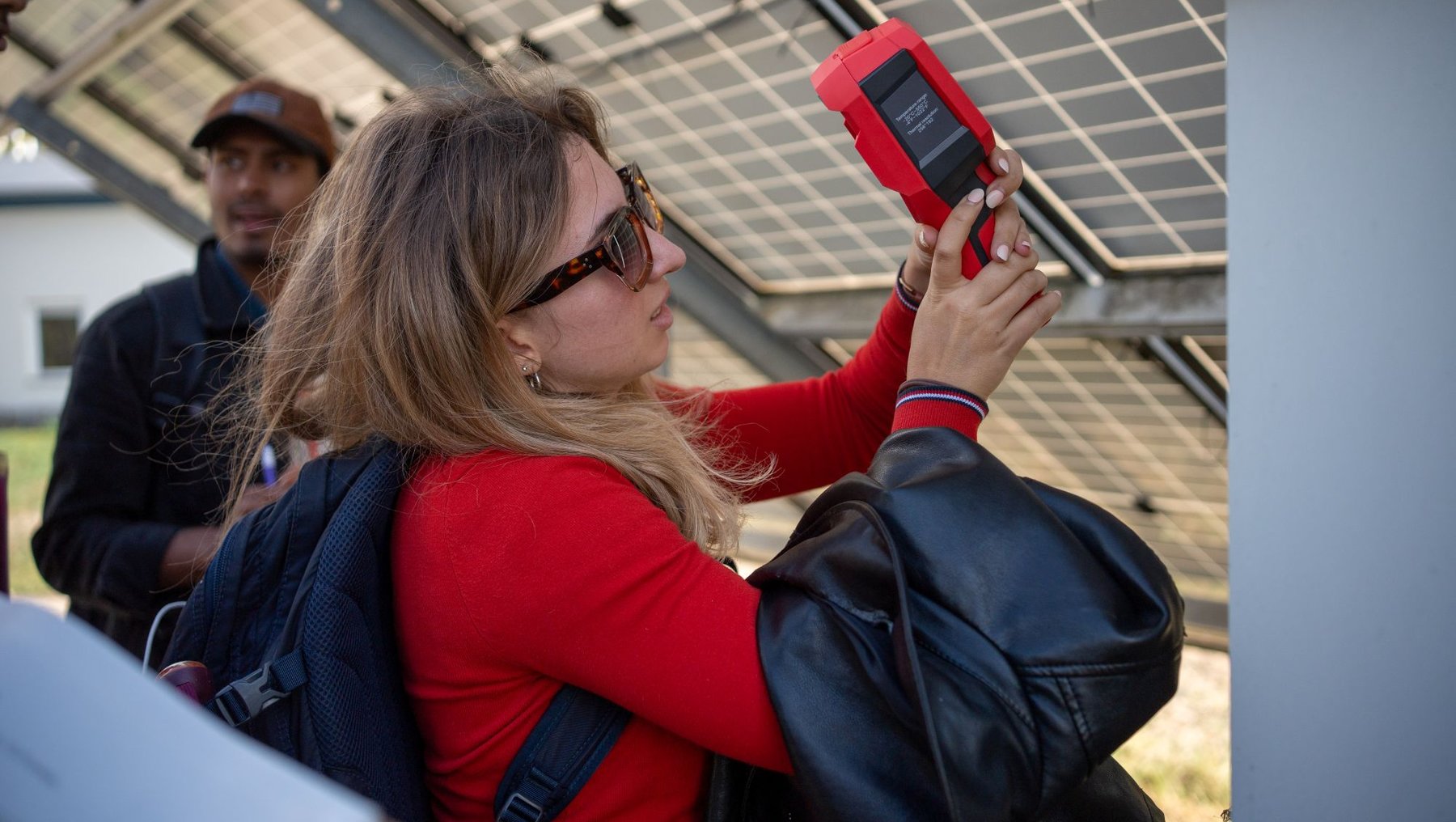 Eine Frau mit Sonnenbrille und rotem Shirt hält ein rotes Meesgerät vor ein Solarpanel und betrachtet es aufmerksam, während im Hintergrund weitere Personen und Agri-PV-Module zu sehen sind.