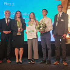 Drei Personen auf einer Bühne erhalten den dritten Platz beim Hugo-Junkers-Preis für Forschung und Innovation aus Sachsen-Anhalt. Die Preisträger halten eine Urkunde und einen Blumenstrauß. Zwei Begleitpersonen stehen daneben.