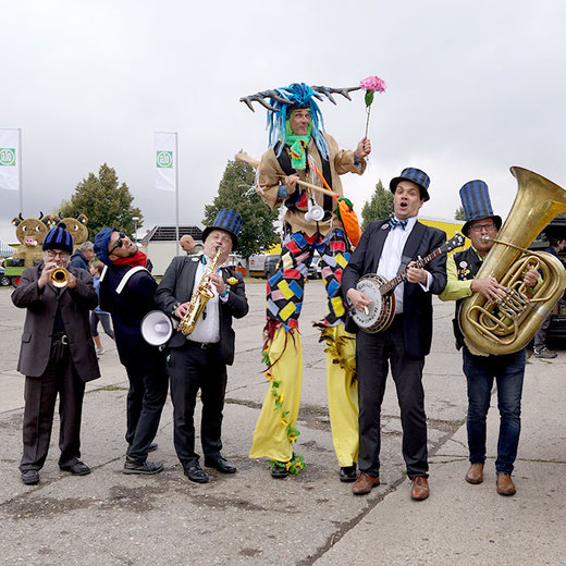 A group of musicians in whimsical costumes, including a person in a colorful, elaborate outfit holding a flower, performing outdoors. They play various instruments like trombones, a banjo, and a tuba, creating a lively and festive atmosphere.