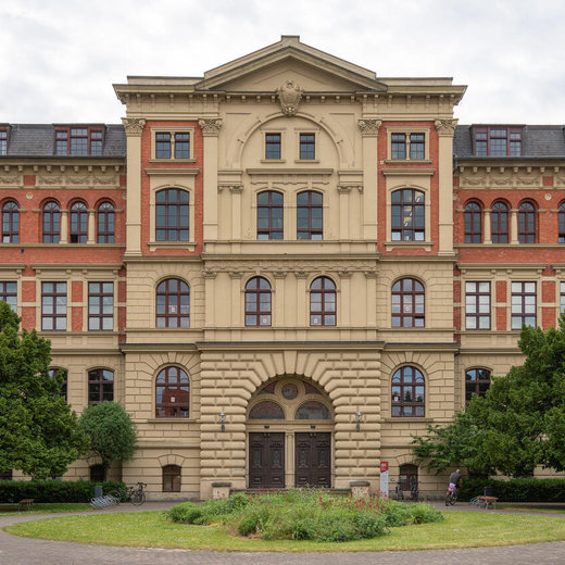 Prächtiges, historisches Gebäude mit klassizistischer Fassade aus rotem Backstein und hellem Sandstein, zentraler Eingang mit Säulen und Rundbogen, drei Stockwerke mit großen, symmetrisch angeordneten Fenstern, umgeben von grünem Rasen und Bäumen.