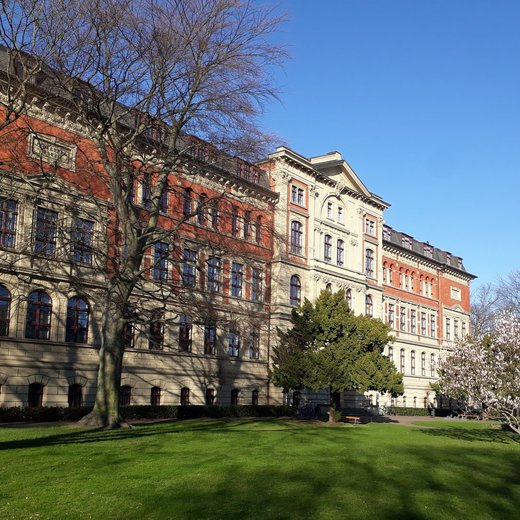 Großes, historisches Gebäude mit rotem Backstein und klassizistischer Architektur, umgeben von kahlen Bäumen und einem gepflegten Rasen vor hellem Himmel.