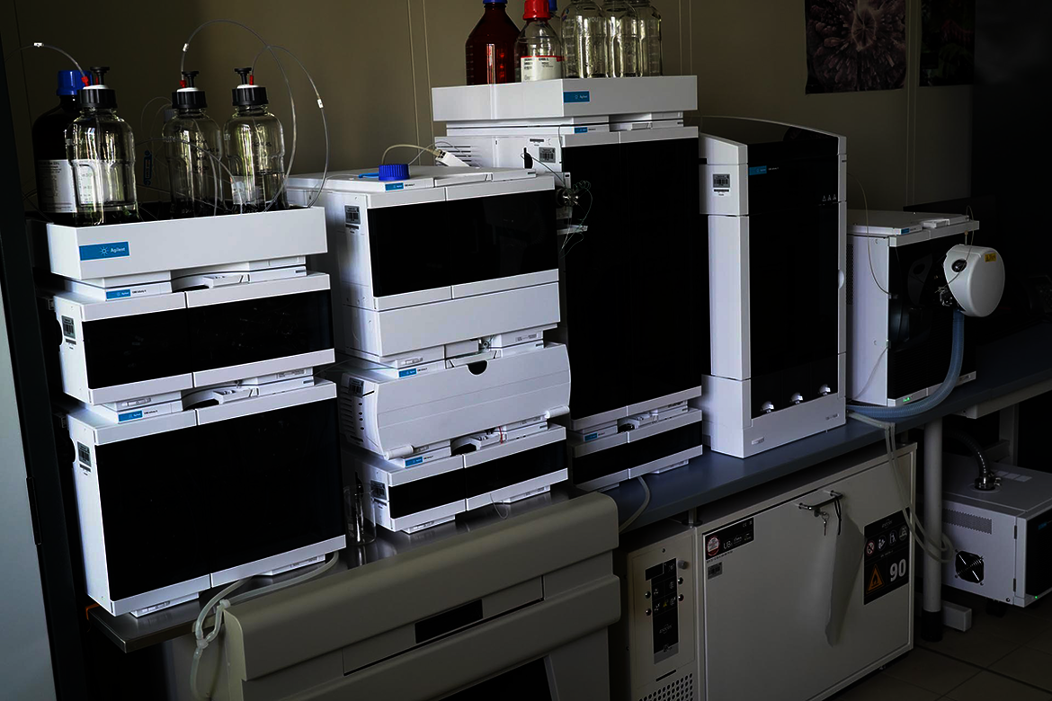 Laborplatz mit zwei großen analytischen Geräten, vermutlich Gaschromatographen oder Flüssigkeitschromatographen, auf einem Tisch. Daneben stehen Flüssigkeitsbehälter und eine Wasserstation. Arbeitsumgebung mit Laborausstattung.