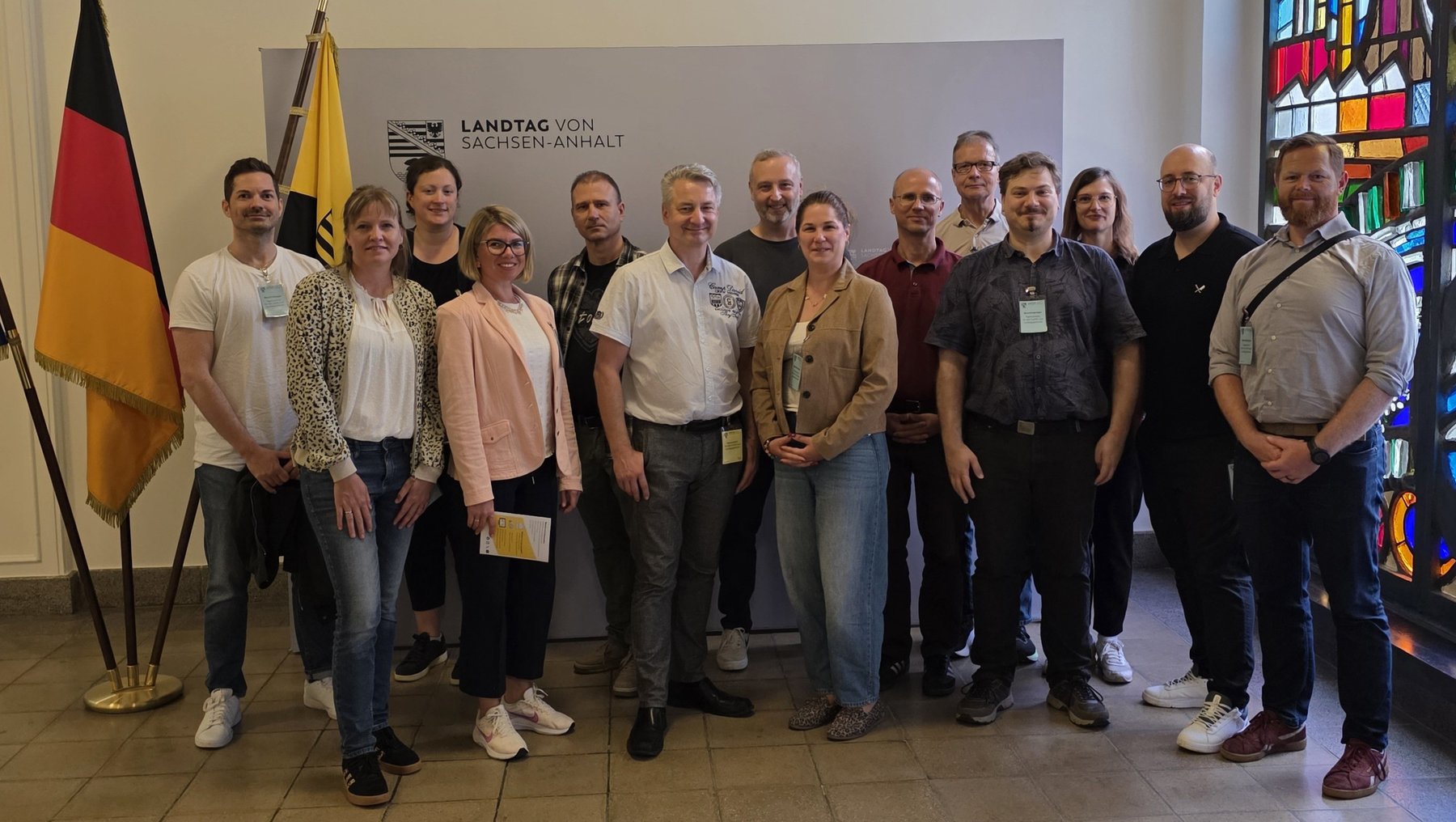 Gruppe von vierzehn Personen steht lächelnd für Gruppenfoto zusammen, darunter Männer und Frauen in Business-Casual-Kleidung. Im Hintergrund deutsche Flagge sowie ein Banner mit dem Text „Landtag von Sachsen-Anhalt“.