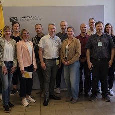Gruppe von vierzehn Personen steht lächelnd für Gruppenfoto zusammen, darunter Männer und Frauen in Business-Casual-Kleidung. Im Hintergrund deutsche Flagge sowie ein Banner mit dem Text „Landtag von Sachsen-Anhalt“.