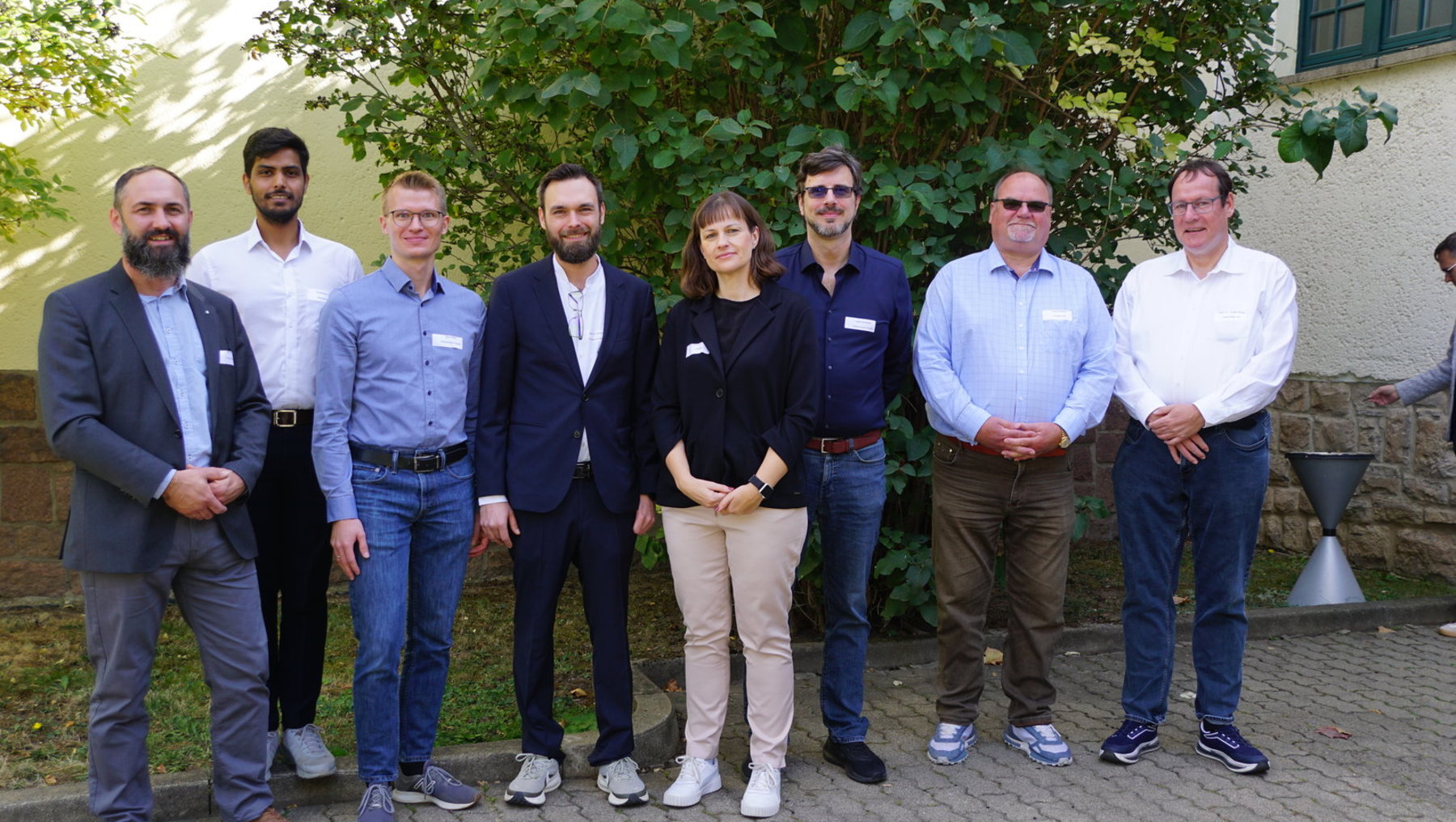Gruppe von sieben Personen steht im Freien vor einem Gebäude mit Bäumen im Hintergrund. Alle tragen Namensschilder und lächeln leicht. Die Kleidung variiert von Business-Casual bis zu lockerer Freizeitkleidung.