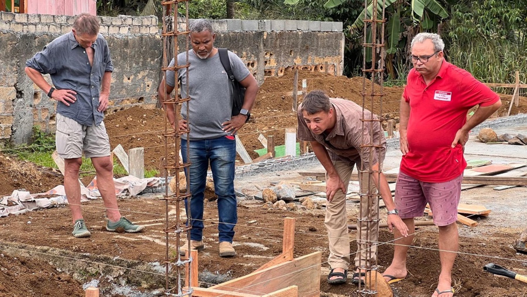 Vier Männer auf einer Baustelle: Drei beobachten und besprechen, während einer in einer Holzform für eine Betonsäule arbeitet, die mit Stahlstangen verstärkt ist.