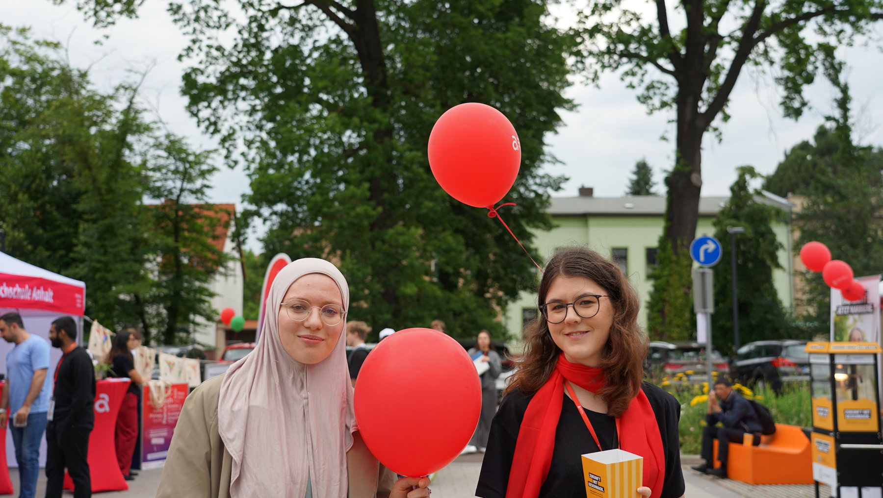 Zwei Personen halten gemeinsam einen roten Luftballon. Die Person links trägt ein helles Kopftuch und ein langes Oberteil, die Person rechts eine Brille, eine rote Schärpe und hält einen Becher. Sie lächeln und wirken fröhlich.