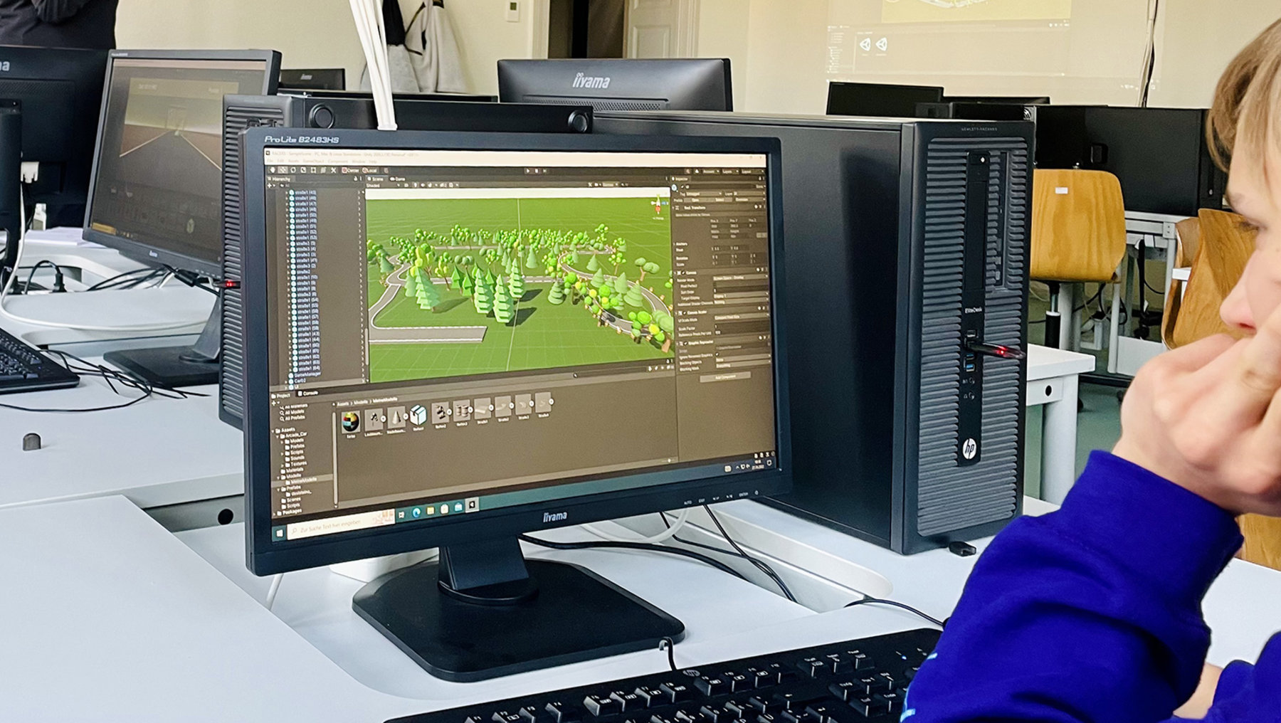 Person nutzt einen Computer mit einer 3D-Software, die ein grünes, begehbares Gelände mit Gebäuden und Wegen anzeigt. Die Person hält eine Maus in der Hand und arbeitet am Bildschirm.