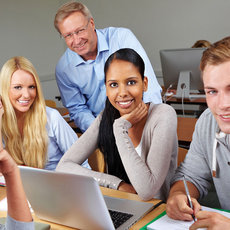Eine Gruppe von sieben lächelnden Erwachsenen sitzt um einen Tisch in einem Klassenzimmer. Sie arbeiten mit Laptops und Notizen. Die Atmosphäre wirkt freundlich und kooperativ, während sie gemeinsam lernen oder eine Besprechung abhalten.