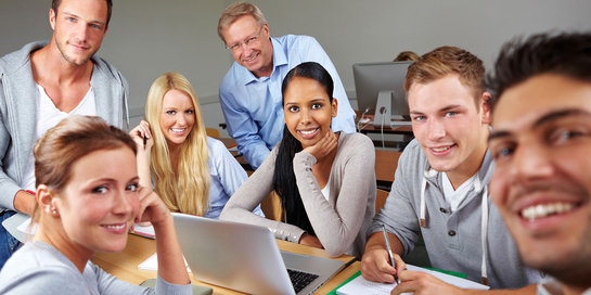 A group of six diverse individuals, both men and women, are sitting around a table in a classroom setting. They are smiling and appear engaged in a discussion or study session. Some are using laptops and notebooks, indicating an academic or collaborative environment.