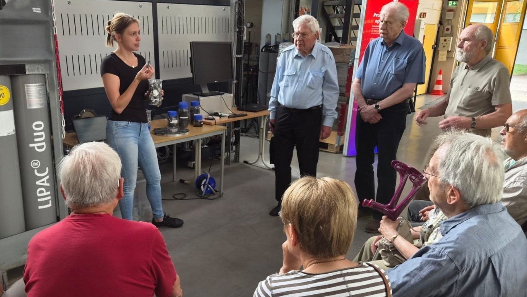 Eine junge Frau erklärt im Industrielabor der Hochschule Anhalt einer Gruppe älterer Menschen, die aufmerksam zuhören und teilweise Notizen machen, die technischen Geräte und Prozesse. Im Hintergrund stehen weitere Personen und Werkzeuge.