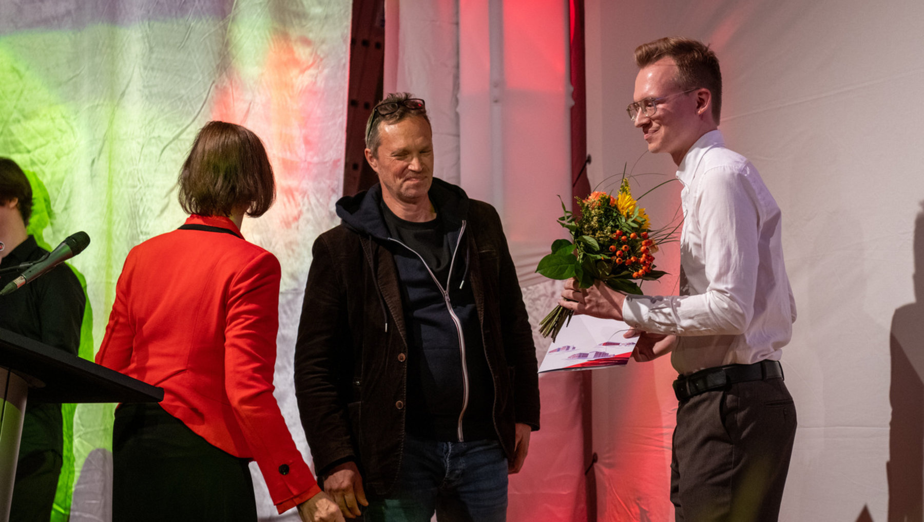 Ein Mann in weißem Hemd überreicht einem anderen Mann mit dunkler Jacke einen Blumenstrauß auf einer Bühne. Eine Frau in rotem Blazer steht daneben und beobachtet die Szene. Der Überreichende hält zusätzlich ein Dokument.