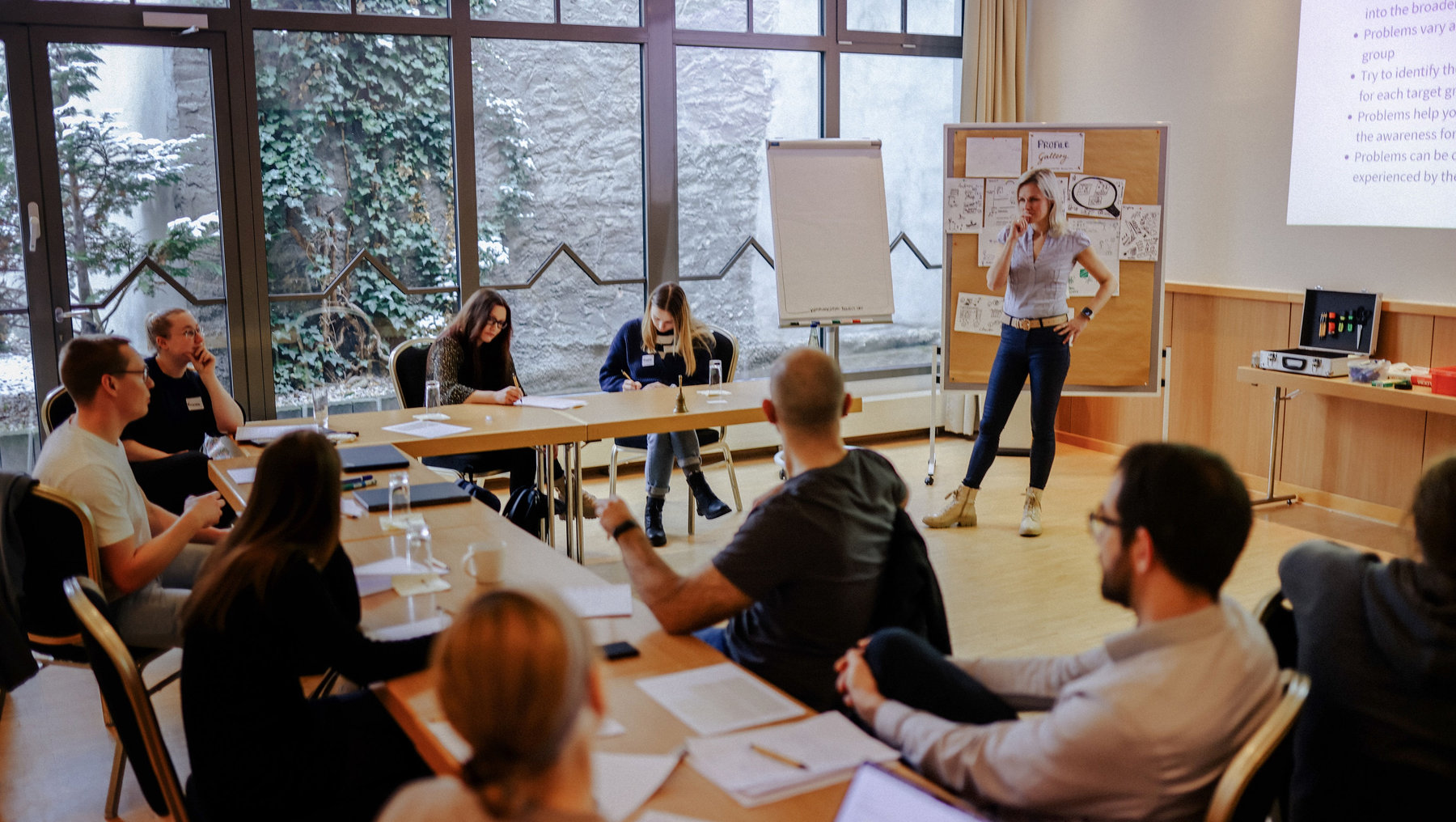 Eine Frau steht vor einer Gruppe von Teilnehmern an einem runden Tisch und hält eine Präsentation zu einem Thema namens „Raise Awareness“. Die Zuhörer sind aufmerksam und notieren sich Punkte.