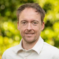 Ein lächelnder Mann mit kurz geschnittenen, hellbraunen Haaren, der ein weißes Hemd trägt und vor einem unscharfen grün-gelben Hintergrund steht.