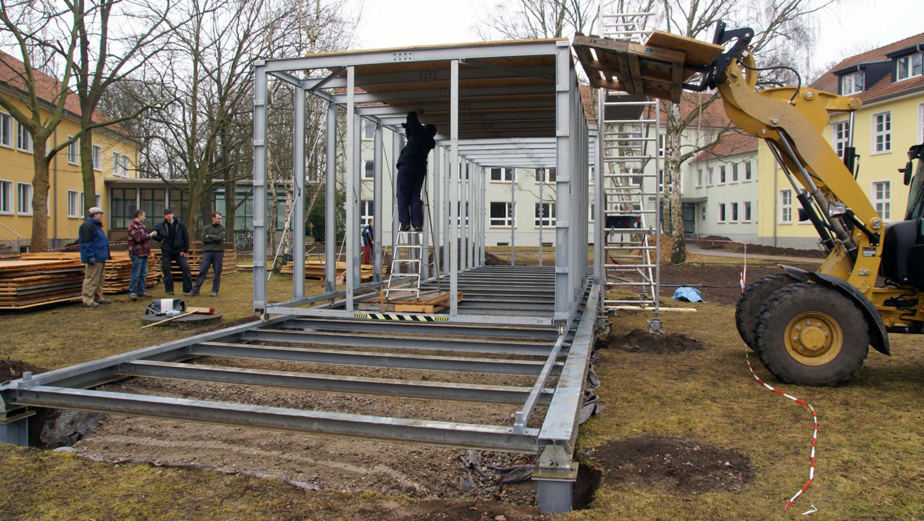 Ein Bauprojekt mit einem teilweise fertiggestellten Holzrahmengebäude. Mehrere Personen stehen um das Gebäude herum und beobachten die Arbeiten. Ein Bagger ist in der Nähe positioniert. Das Gelände ist mit Baumaterialien übersät.