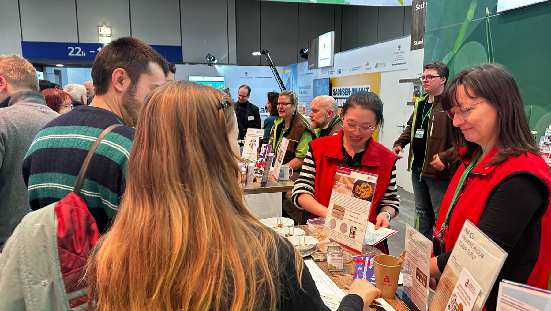 Menschen auf einer Messe stehen an einem Informationsstand mit Prospekten und kleinen Gläsern, während eine Mitarbeiterin in rotem Jackett lächelnd Informationsmaterial aushändigt. Im Hintergrund sind weitere Besucher und ein Stand mit dem Schild „Sachsen-Anhalt“.