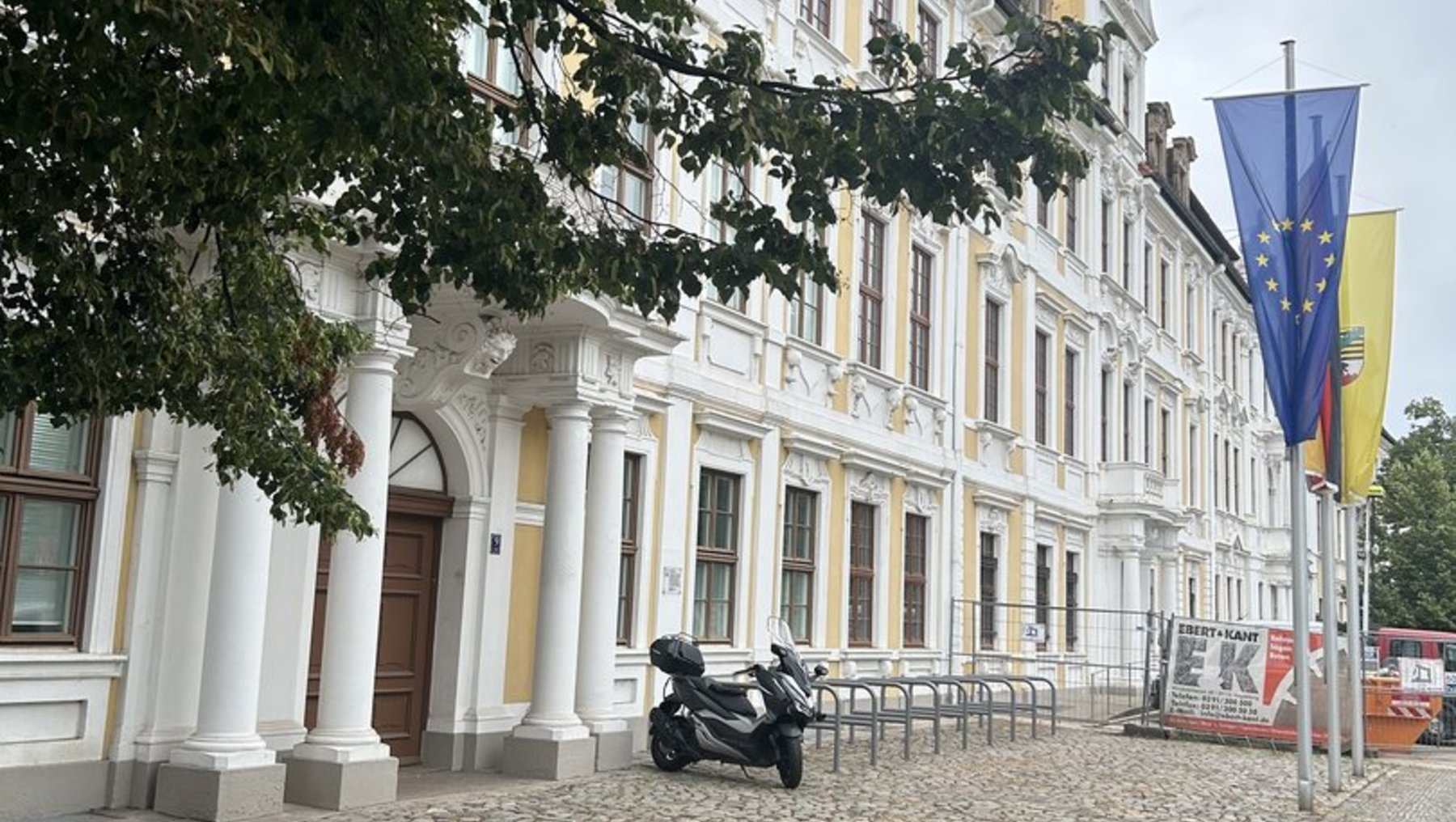 Historisches, weißes Gebäude mit klassizistischer Architektur, Säulen und Balkon. Vor dem Eingang steht eine Motorradstatue. Zwei Flaggenmasten mit der EU-Flagge und einer weiteren Flagge sind vor dem Gebäude aufgestellt. Pflasterstraße mit Baum im Vordergrund.