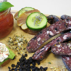 This_image_displays_a_charcuterie_plate_with_sliced_cured_meats_pastries_spread_with_ripe_tomatoes_olives_cucumber_and_a_small_bowl_of_tomato_sauce.

Alternative description: "Assorted charcuterie with meats, spreads, sauces, veggies, and bread slices."