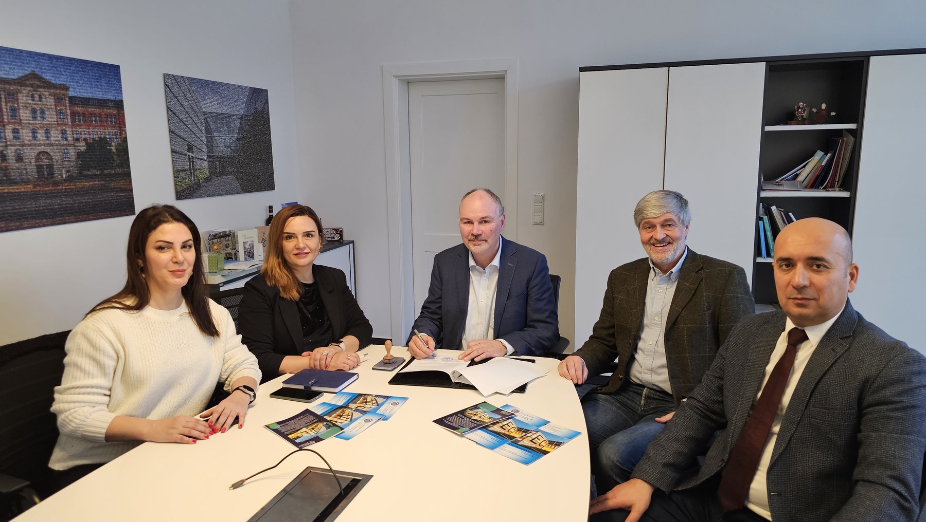 Ein Meeting mit fünf Personen in einem Besprechungsraum. Drei Männer und zwei Frauen sitzen um einen Tisch mit Unterlagen und Broschüren. Die Wände sind mit Bildern geschmückt, und ein Regal mit Büchern ist im Hintergrund zu sehen. Alle wirken konzentriert und professionell.