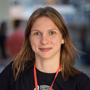 Eine lächelnde Frau mit langen, hellbraunen Haaren, die ein schwarzes Shirt mit bedrucktem Aufdruck trägt und eine rote Schleife oder ein Band um den Hals hat.