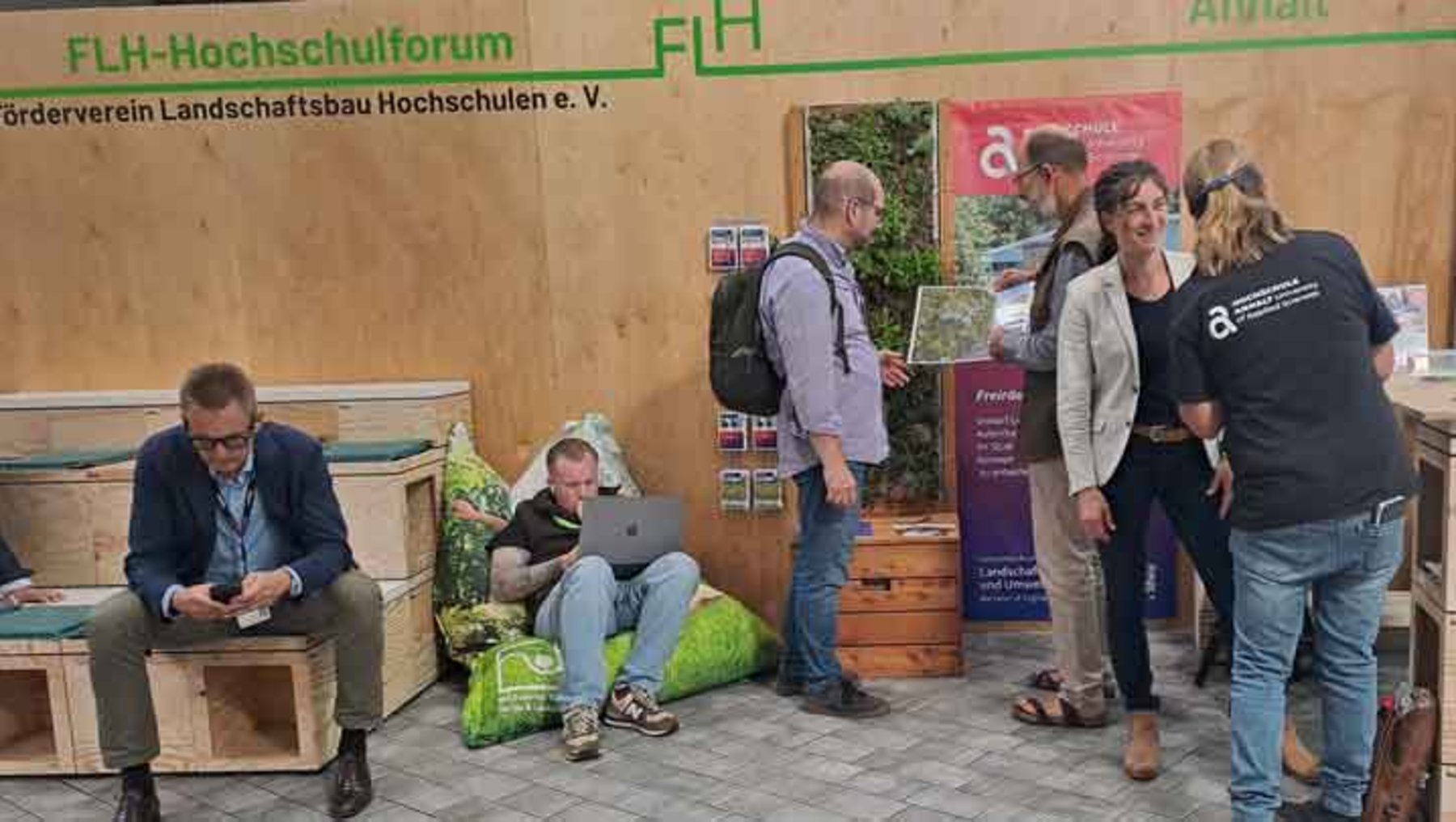 Auf einer Messe stehen mehrere Personen vor einem Infostand des Fördervereins Landschaftsbau Hochschulen. Links sitzt jemand auf Paletten mit Laptop, andere stehen und unterhalten sich. Im Hintergrund hängen Plakate mit grünen Pflanzenmotiven und Logo des Vereins.