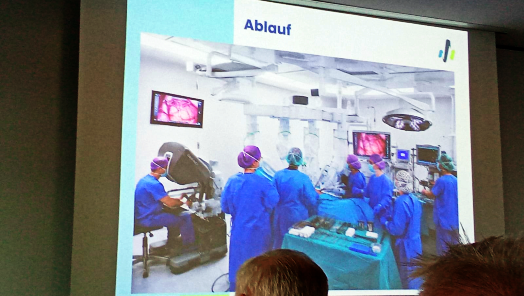 Präsentation zeigt einen Operationssaal mit medizinischem Team in blauer OP-Kleidung, das um einen Patienten mit offenem Brustkorb versammelt ist. Zwei Bildschirme im Hintergrund zeigen eine Laparoskopie. Überschrift lautet „Ablauf“.