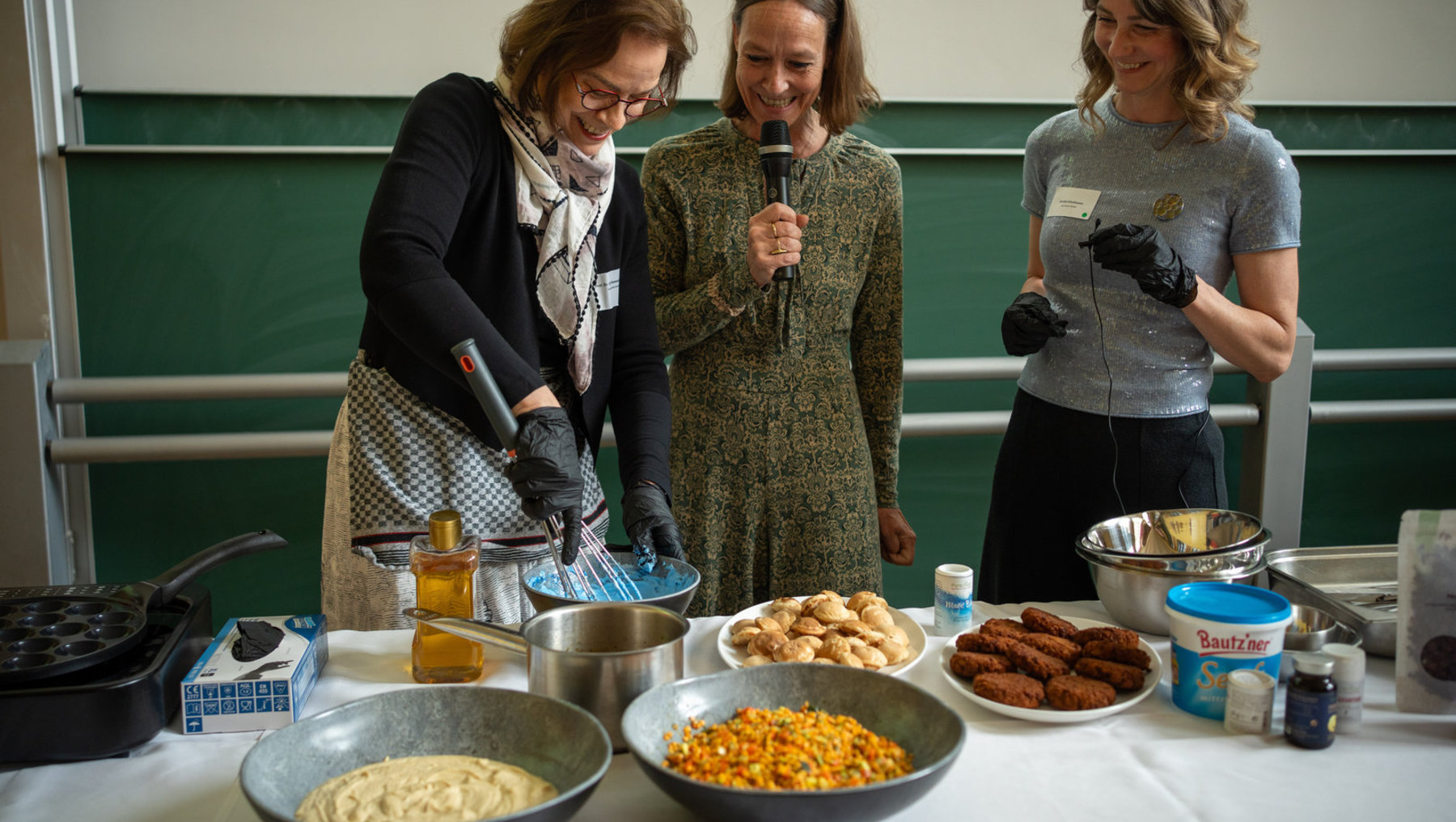 Drei Frauen stehen lächelnd in einer Küche und bereiten gemeinsam Speisen zu. Auf dem Tisch liegen Zutaten wie Teig, Gewürze, fertige Gebäckstücke und eine Packung Bautzner-Milchprodukte. Eine hält ein Mikrofon, eine andere einen Küchenhandschuh.