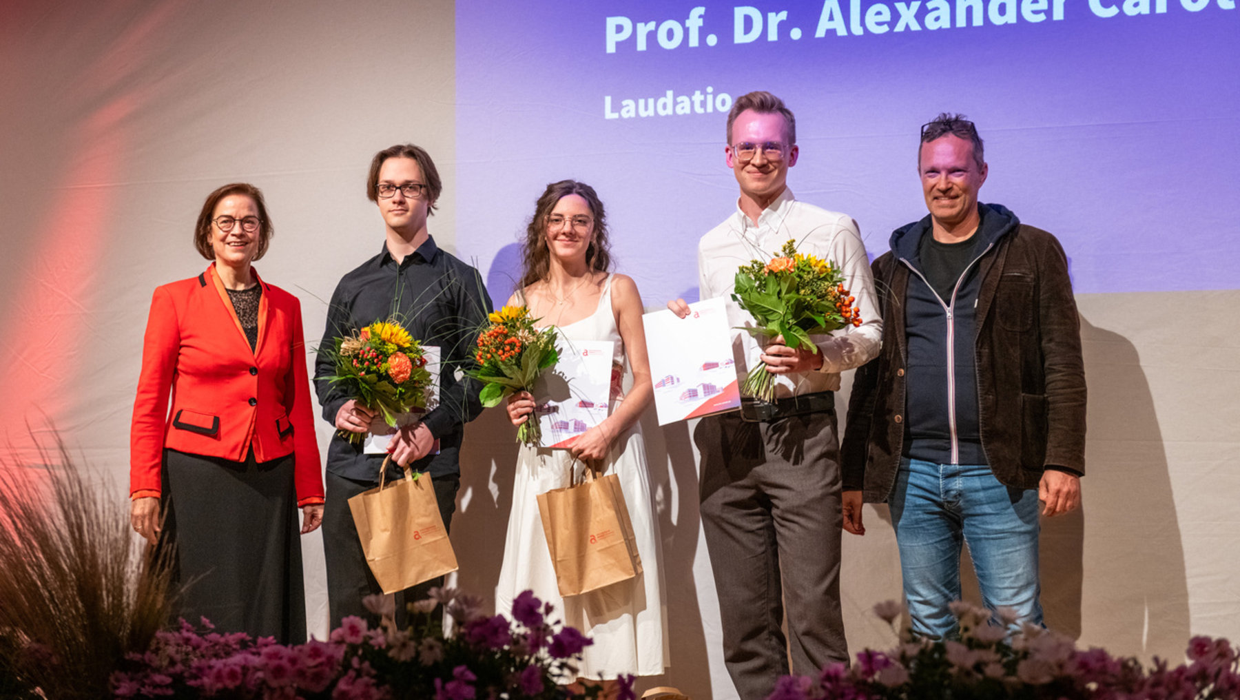 Fünf Personen stehen auf einer Bühne bei einer Preisverleihung. Vier von ihnen halten Blumensträuße und Tüten. Eine Frau in rotem Blazer steht links, gefolgt von drei jüngeren Personen in der Mitte und einem älteren Mann rechts. Im Hintergrund ist der Name Prof. Dr. Alexander Carôt mit Laudatio zu sehen.
