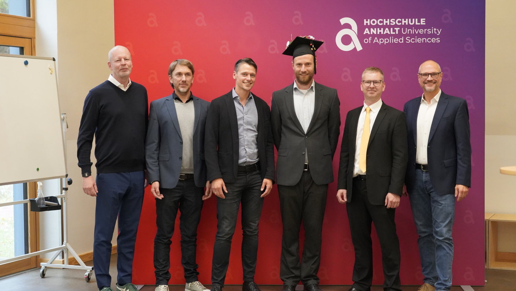 Sechs Männer stehen vor einem roten Hintergrund mit dem Logo der Hochschule Anhalt. Der Mann in der Mitte trägt einen Anzug und einen akademischen Hut, die anderen fünf tragen Anzüge oder Business-Kleidung. Feierlicher Anlass, vermutlich eine Abschlussveranstaltung.