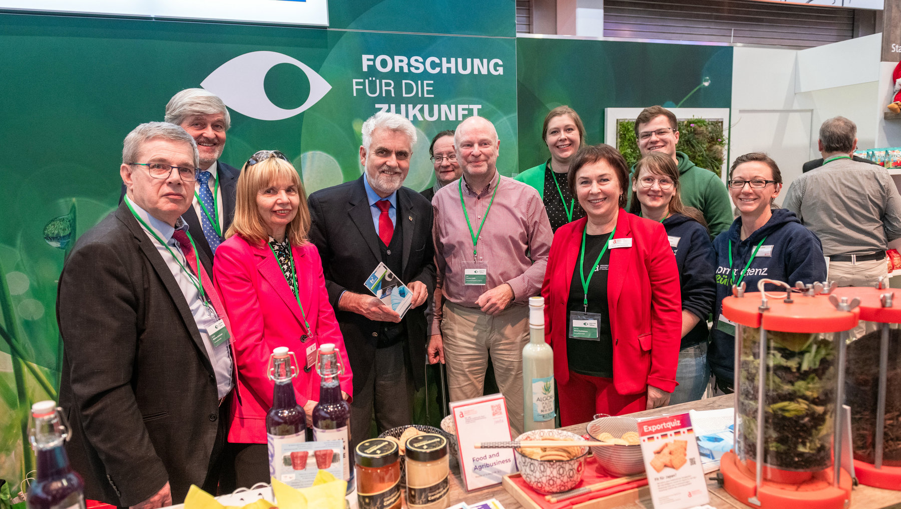Gruppe von Personen vor einem Messestand mit dem Motto „Forschung für die Zukunft“. Sie stehen vor verschiedenen Produkten wie Getränken, Gläsern mit Algen und Informationsmaterialien zu Lebensmitteln und Agrarwirtschaft.