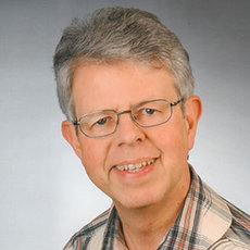 A middle-aged man with short gray hair, wearing glasses and a plaid shirt, smiling at the camera. The background is plain and neutral, focusing attention on the man's face.