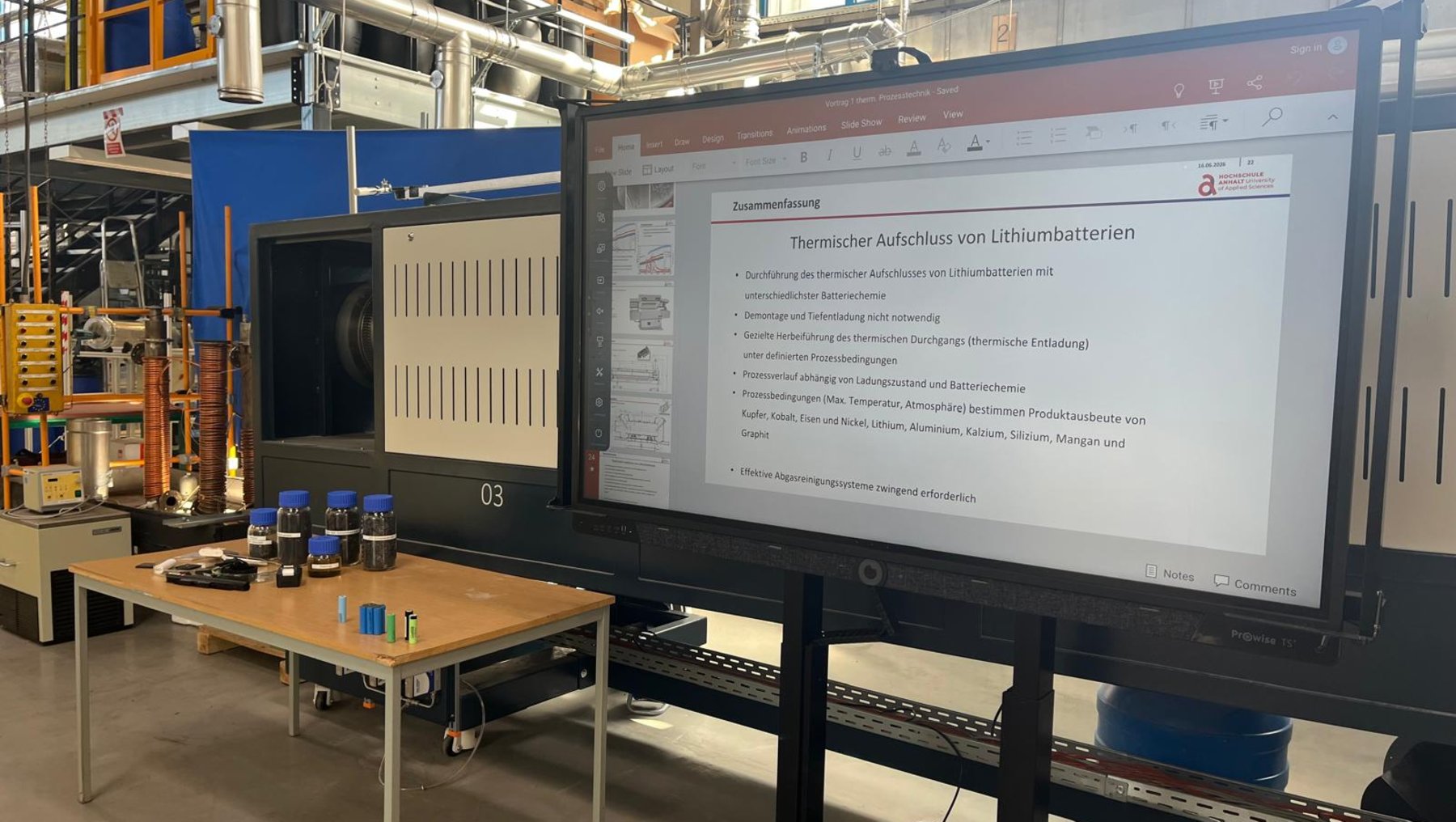 Präsentation in einer Produktionshalle mit dem Titel „Thermischer Aufschluss von Lithiumbatterien“. Gezeigt werden eine große Maschine zur Batterieverarbeitung sowie eine Liste mit Schritten wie Zerkleinern, Sortieren und chemischer Extraktion von Lithium, Kobalt und anderen Metallen.