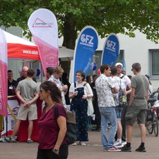 Menschenmenge auf einer Messe oder einem Event, die sich vor mehreren Infoständen mit Flaggen und Werbebannern versammelt, darunter die Marken „Vertam CEO“ und „fz“. Die Besucher stehen in lockerer Gesprächsrunde, einige halten Getränke oder Unterlagen.