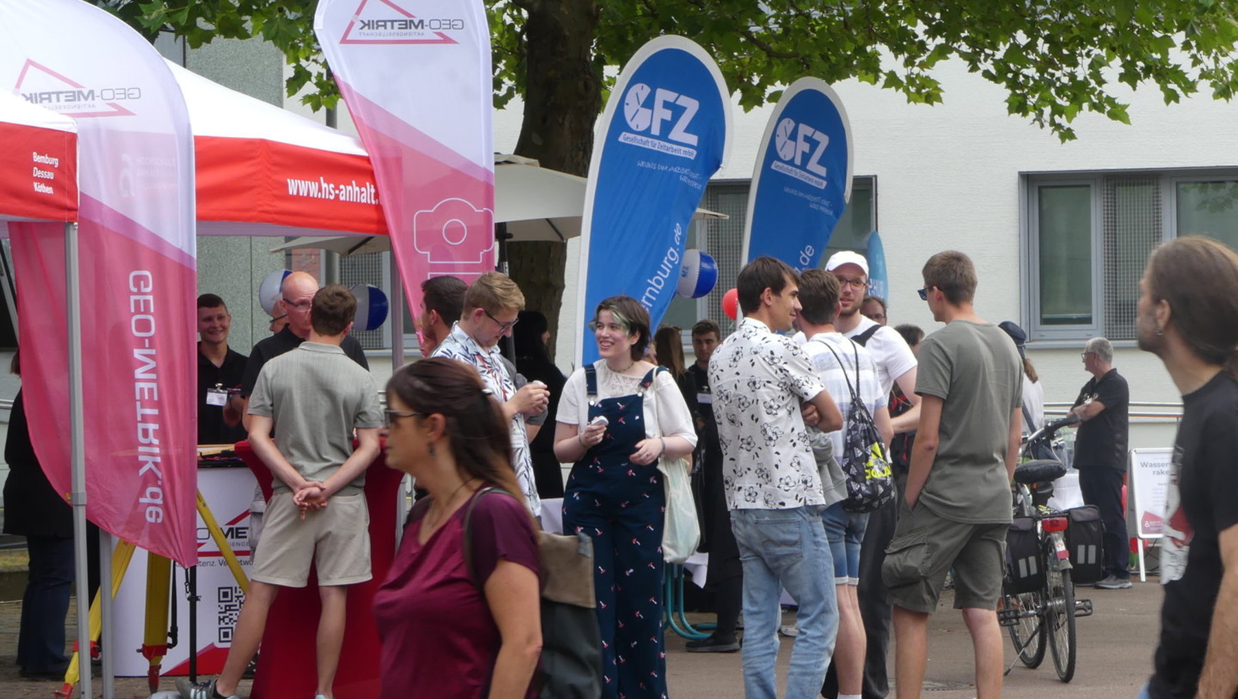 Menschenmenge auf einer Messe oder einem Event, die sich vor mehreren Infoständen mit Flaggen und Werbebannern versammelt, darunter die Marken „Vertam CEO“ und „fz“. Die Besucher stehen in lockerer Gesprächsrunde, einige halten Getränke oder Unterlagen.