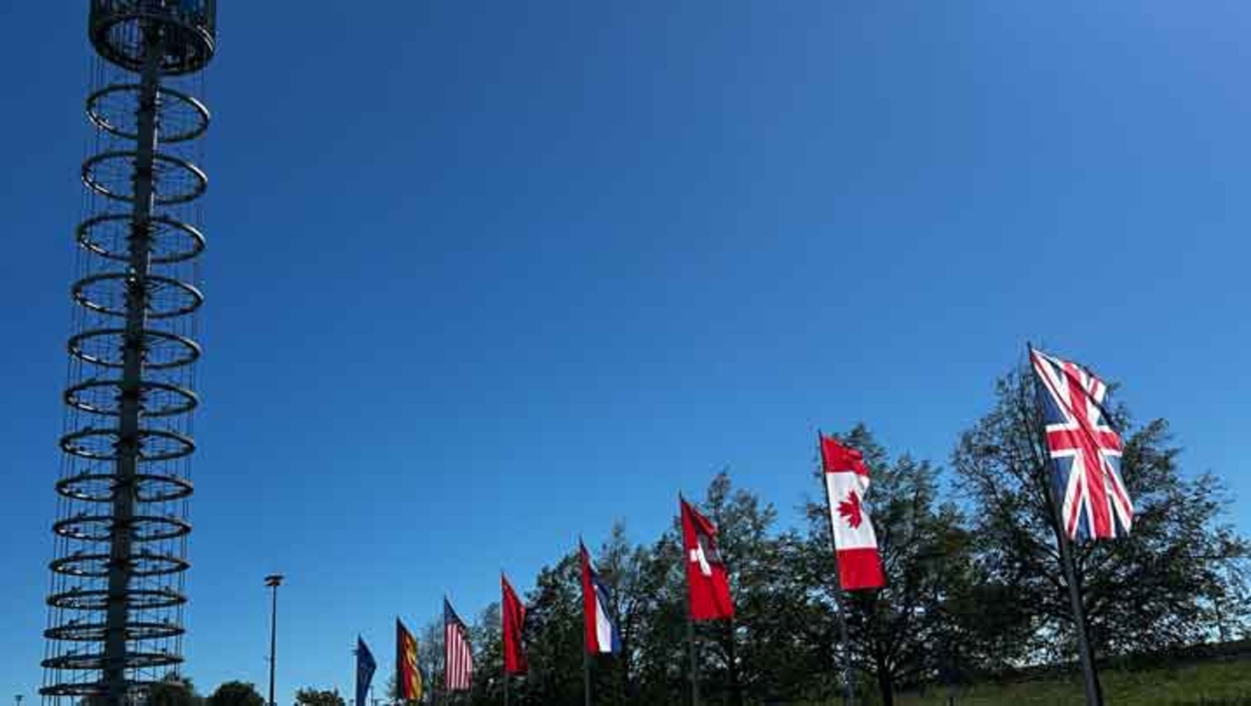 Vor einem klaren Himmel stehen mehrere Flaggen in einer Reihe, darunter die Flaggen Kanadas mit rotem Hintergrund und rotem Ahornblatt sowie die Flagge Großbritanniens mit rotem Kreuz auf weißem Grund. Links im Bild ist ein hoher Sendemast zu sehen.